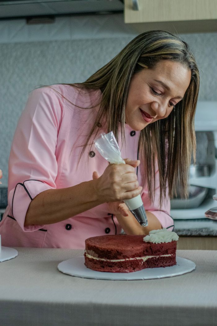 Woman in a pink chef jacket skillfully decorates a cake with frosting in a kitchen setting.