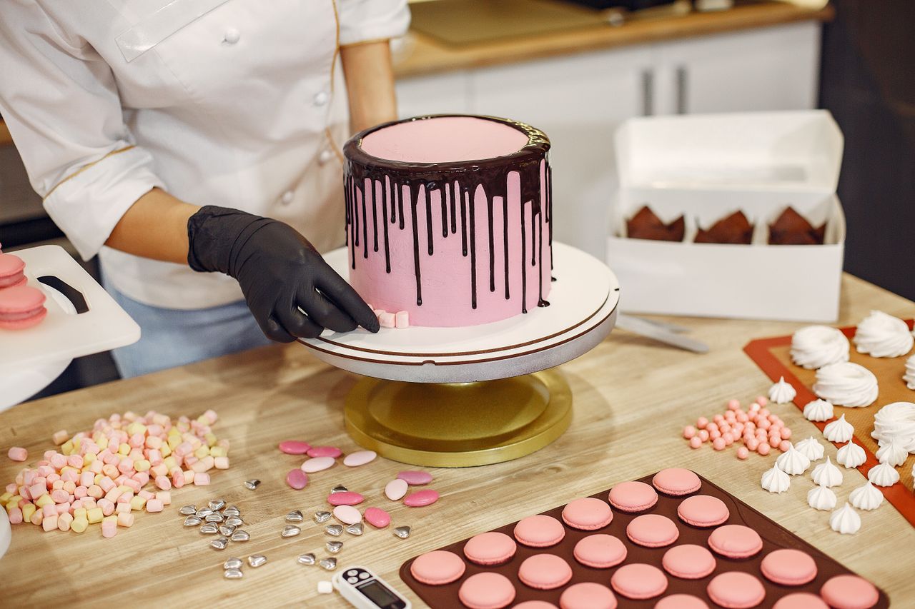 From above crop female cook in uniform and latex gloves placing small sweets on bottom of cake placed on stand near desserts in confectionery